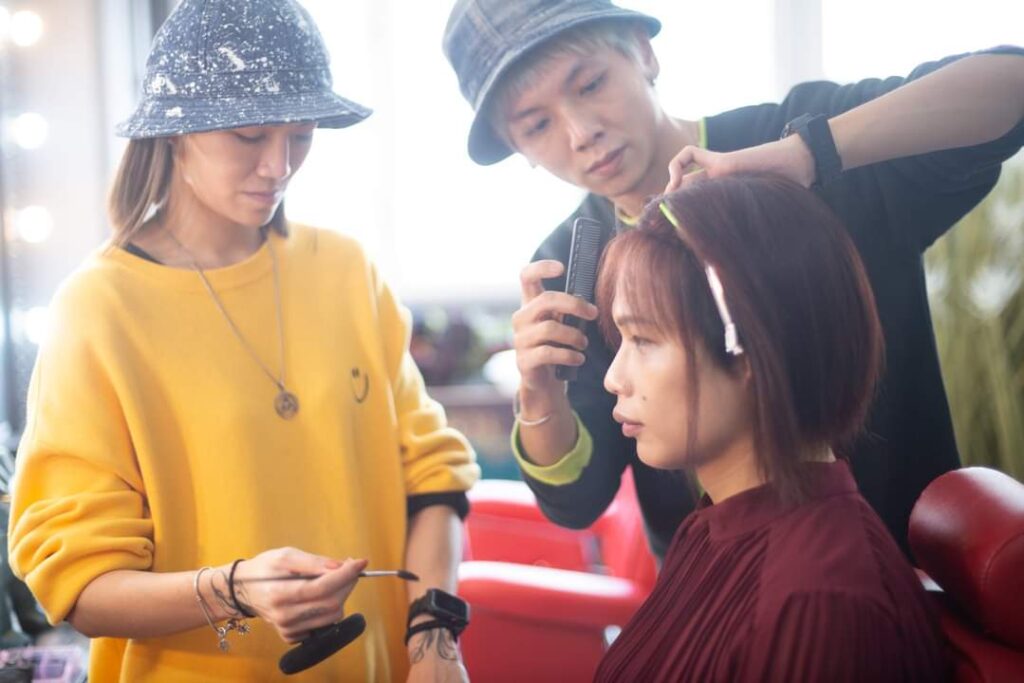 A person with short auburn hair sits in a red chair while two people in a yellow sweater and a black top style their hair and makeup.